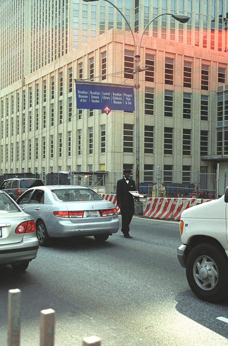 Foto uit New York (2003) van een krantenverkoper in pak die tussen het verkeer op straat loopt.