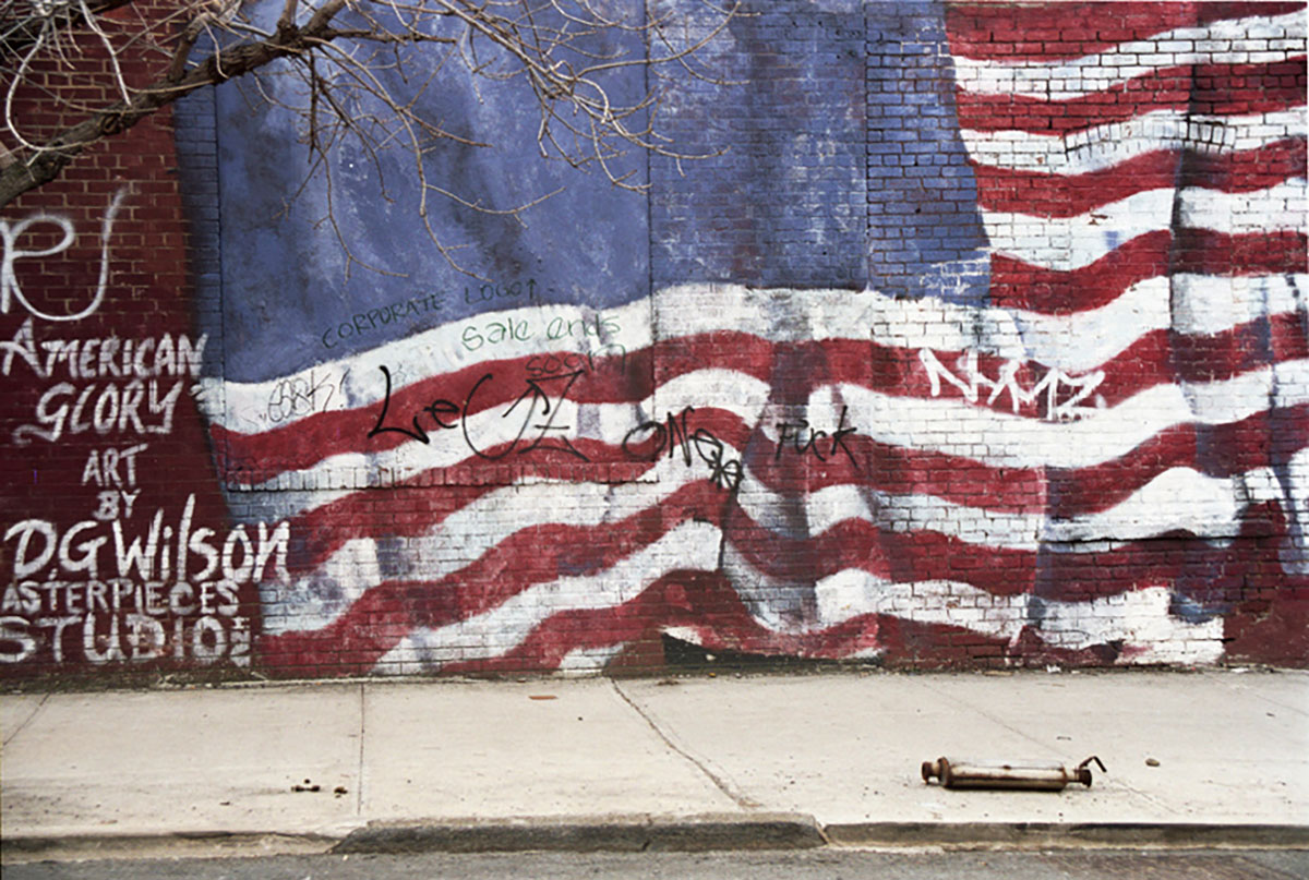 Foto uit New York (2003) van een muurschildering American Glory van een Amerikaanse vlag op een bakstenen muur, overdekt met graffiti, met een uitlaat op de stoep ervoor.