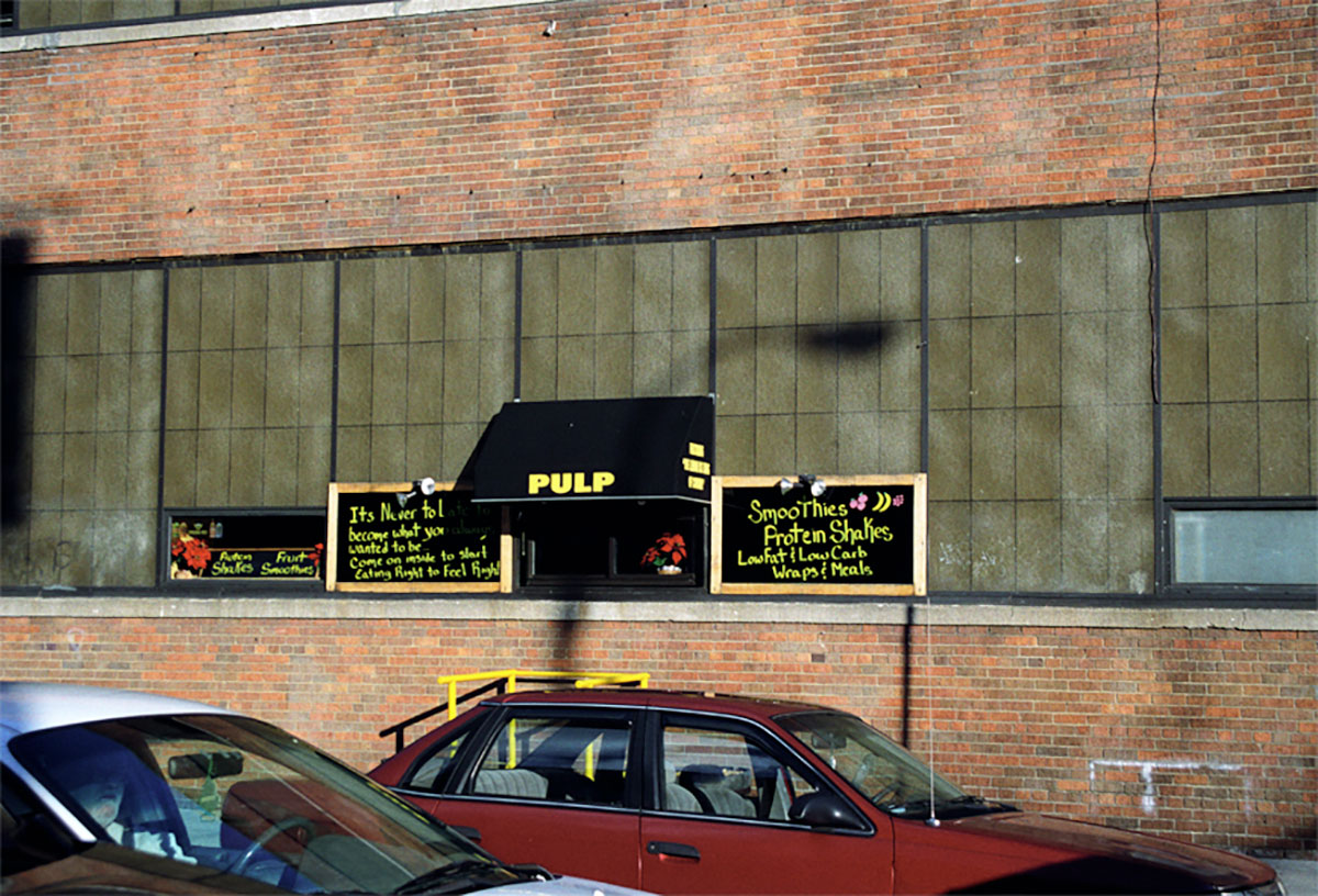 Foto uit New York (2003) van de gevel van smoothiebar Pulp, met met de hand beschreven borden, achter geparkeerde auto's.