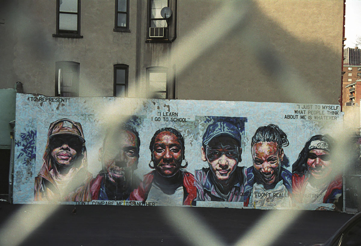 Foto uit New York (2003) van een muurschildering met zes portretten en persoonlijke uitspraken, gefotografeerd door een hek van gaas.