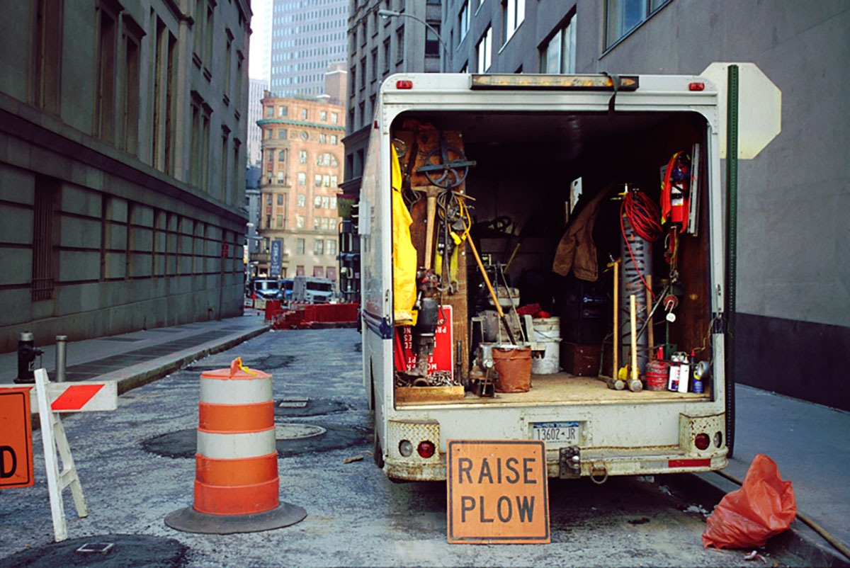 Foto uit New York (2003) van een geopende bestelwagen vol gereedschap, met wegwerkmateriaal ervoor in een straat met hoge gebouwen op de achtergrond.