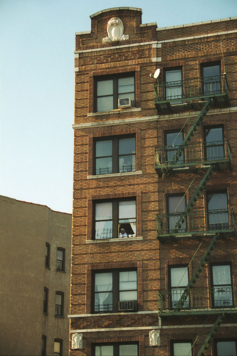 Foto uit New York (2003) van een bakstenen appartementengebouw in Williamsburg, Brooklyn, met brandtrappen langs de gevel en iemand zichtbaar in een van de vensters.