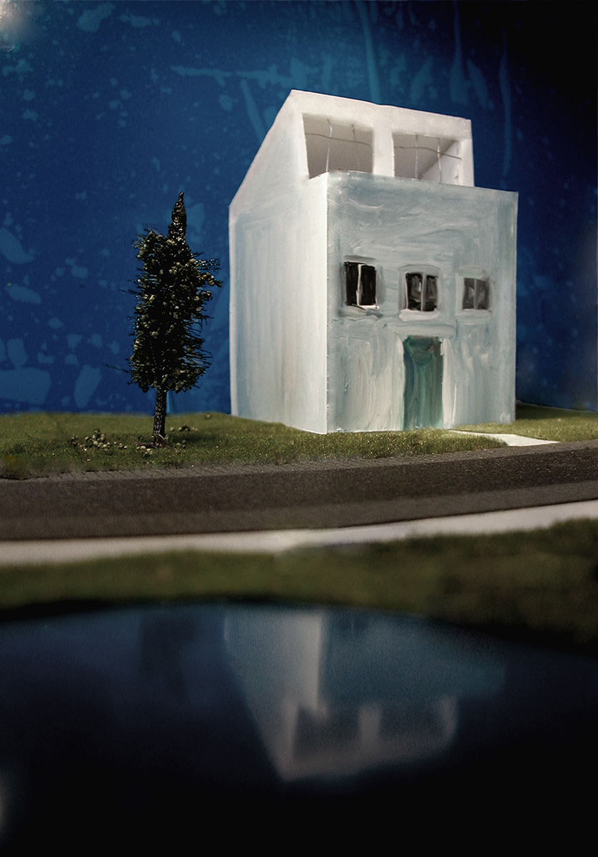 Mixed media werk 'Apocalyps 1': een wit geschilderd maquettehuis naast een boom, aan een weg met een waterplas op de voorgrond, tegen een blauwe achtergrond.