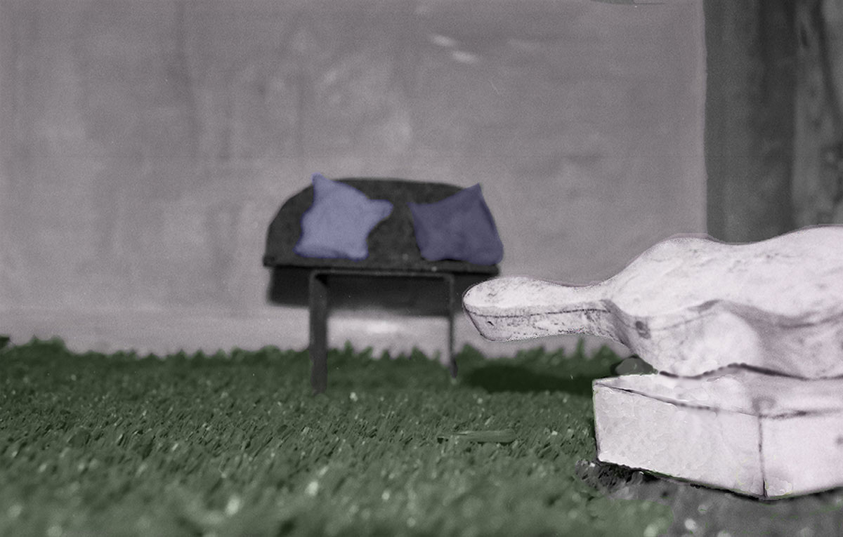 Mixed media werk 'Fantastisch landschap 1': een miniatuurbankje met kussens op kunstgras, met op de voorgrond een wit gipsachtig object.